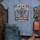 Plumber T-Shirt — "Pipe Doctor. No Appointment Needed" Vintage Work Shirt — blue-jean — Flat-lay Plumber T-Shirt — "Pipe Doctor. No Appointment Needed" Vintage Work Shirt — blue-jean t-shirt, flat-lay product photo | Pressure Tested Co