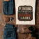 Plumbing Warning Label T-Shirt — Funny Plumber Shirt — natural t-shirt, flat-lay product photo | Pressure Tested Co