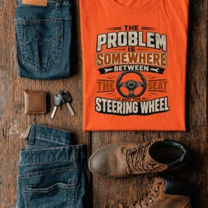 Driving Humor T-Shirt — "The Problem Is Somewhere Between The Seat And The Steering Wheel" — orange t-shirt, flat-lay product photo | Pressure Tested Co